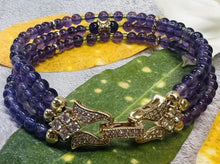 Load image into Gallery viewer, Elegant triple band Amethyst bracelet featuring lustrous purple 6mm and 4mm beads with delicate 18K gold accents and a central gold charm, displayed on green leaves and white petals.